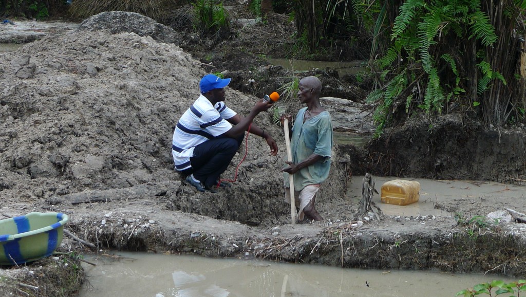 journalist holds microphone up to a miner holding a shovel and standing in a pool of dirty water