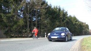 Michael Wigge mit dem Tretroller unterwegs auf einer Landstraße, neben ihm fährt ein Auto